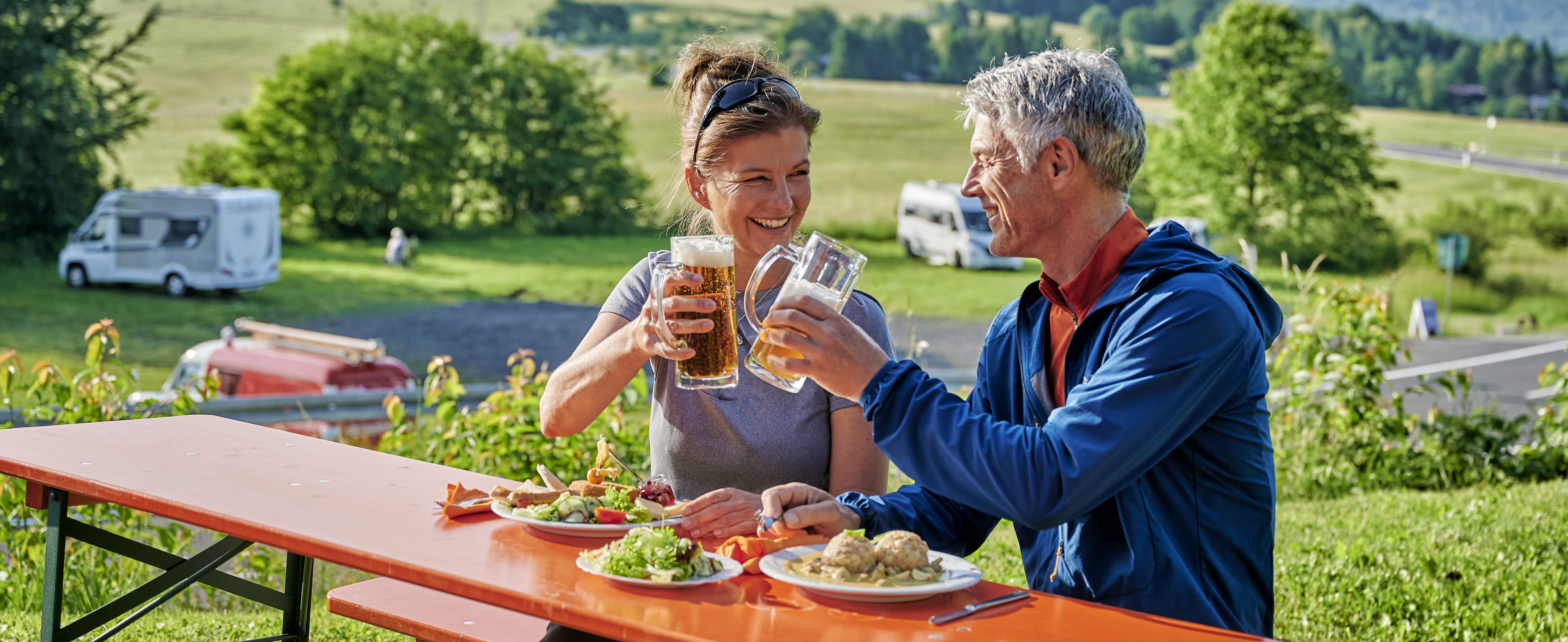 Zwei Personern sitzen an einer Bierzeltgarnitur, haben Teller mit Essen vor sich und prosten sich mit Biergläsern zu.