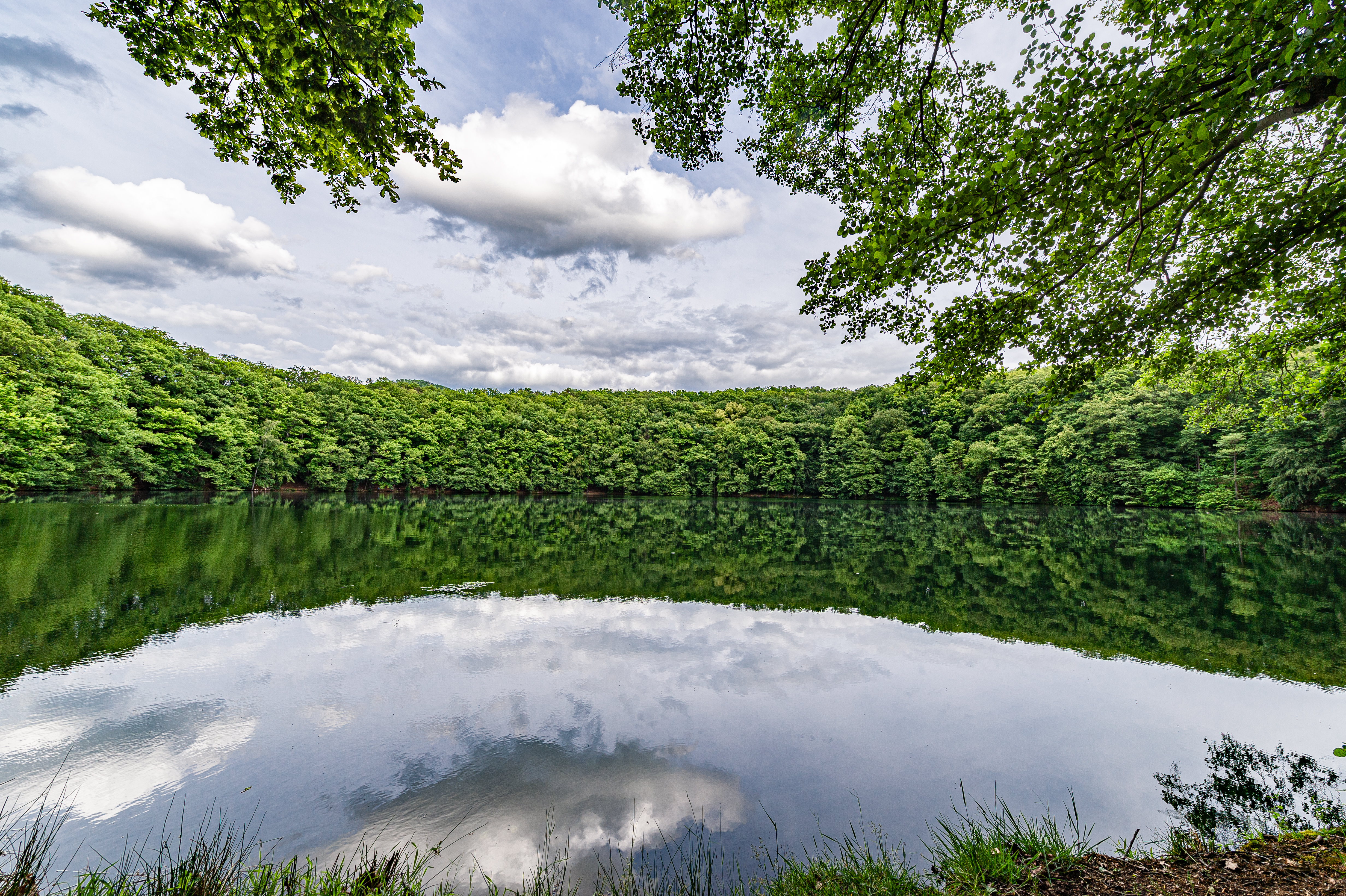 See mit grünen Bäumen ringesherum und Wolkenhimmel, der sich auf der Wasseroberfläche spiegelt.