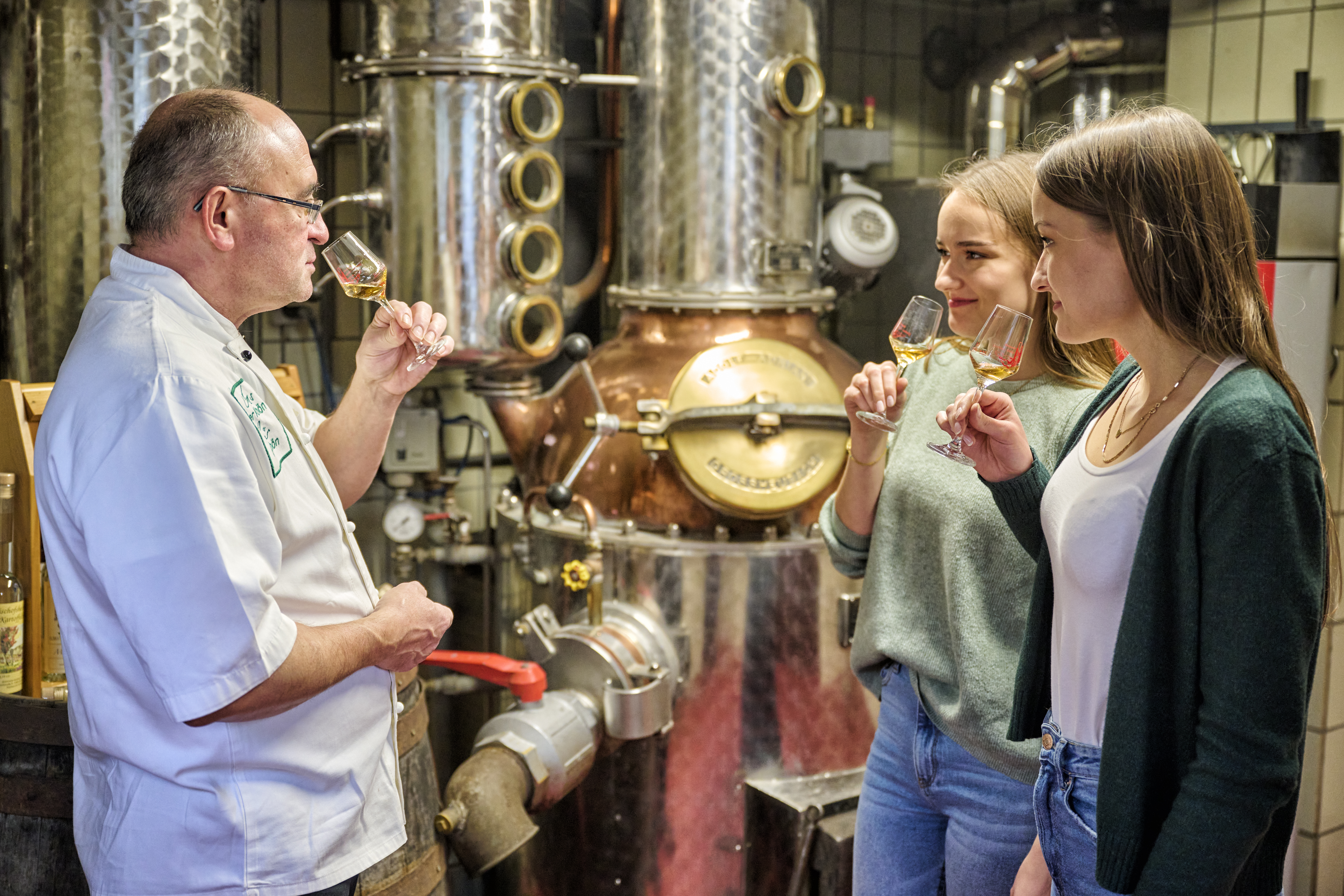 Eine Brennereiführung, bei der zwei Frauen am Glas riechen. Ihnen gegenüber steht der Brenner und riecht ebenfalls am Glas.
