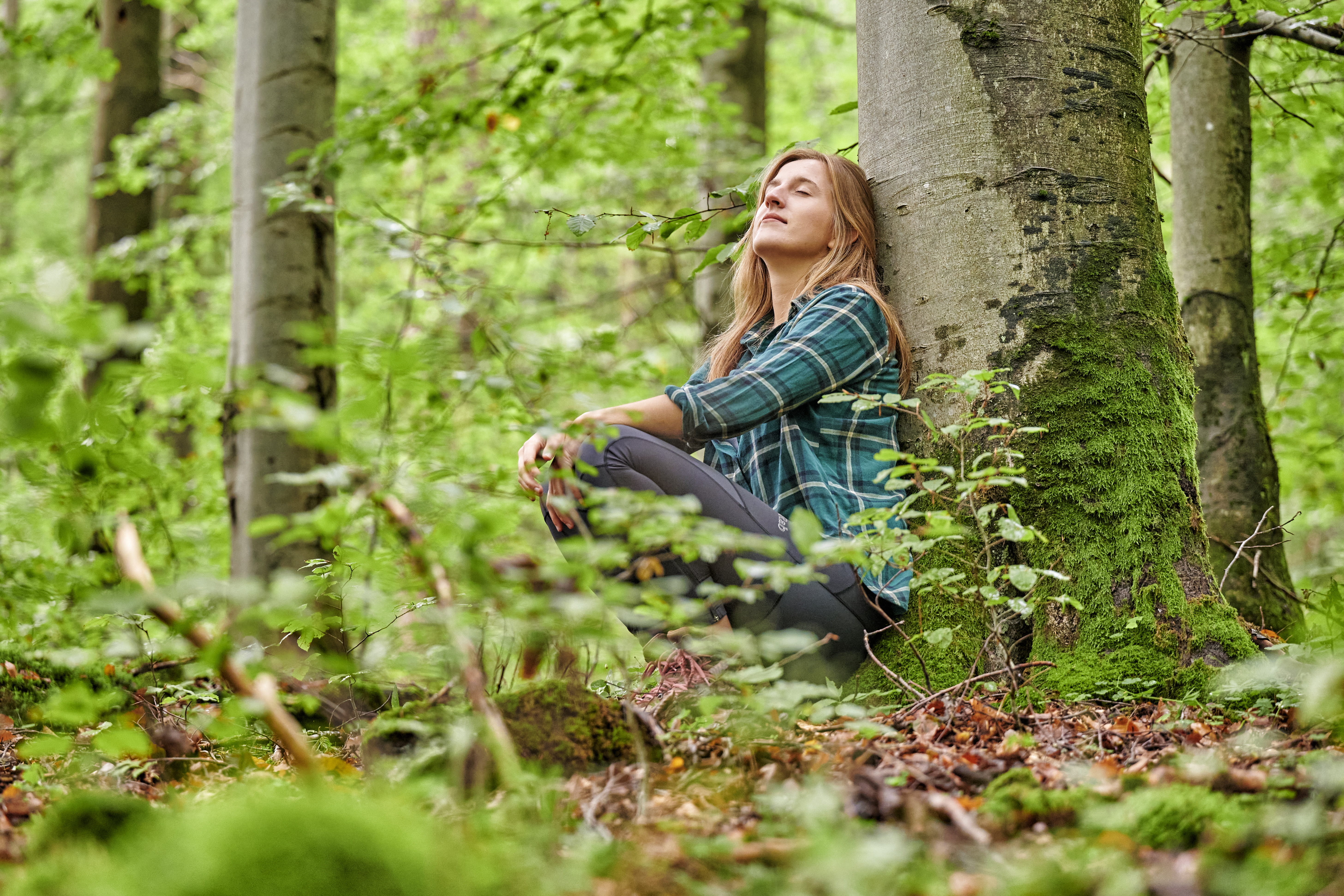 Frau hockt im Wald und lehnt sich entspannt an einem Baum an.