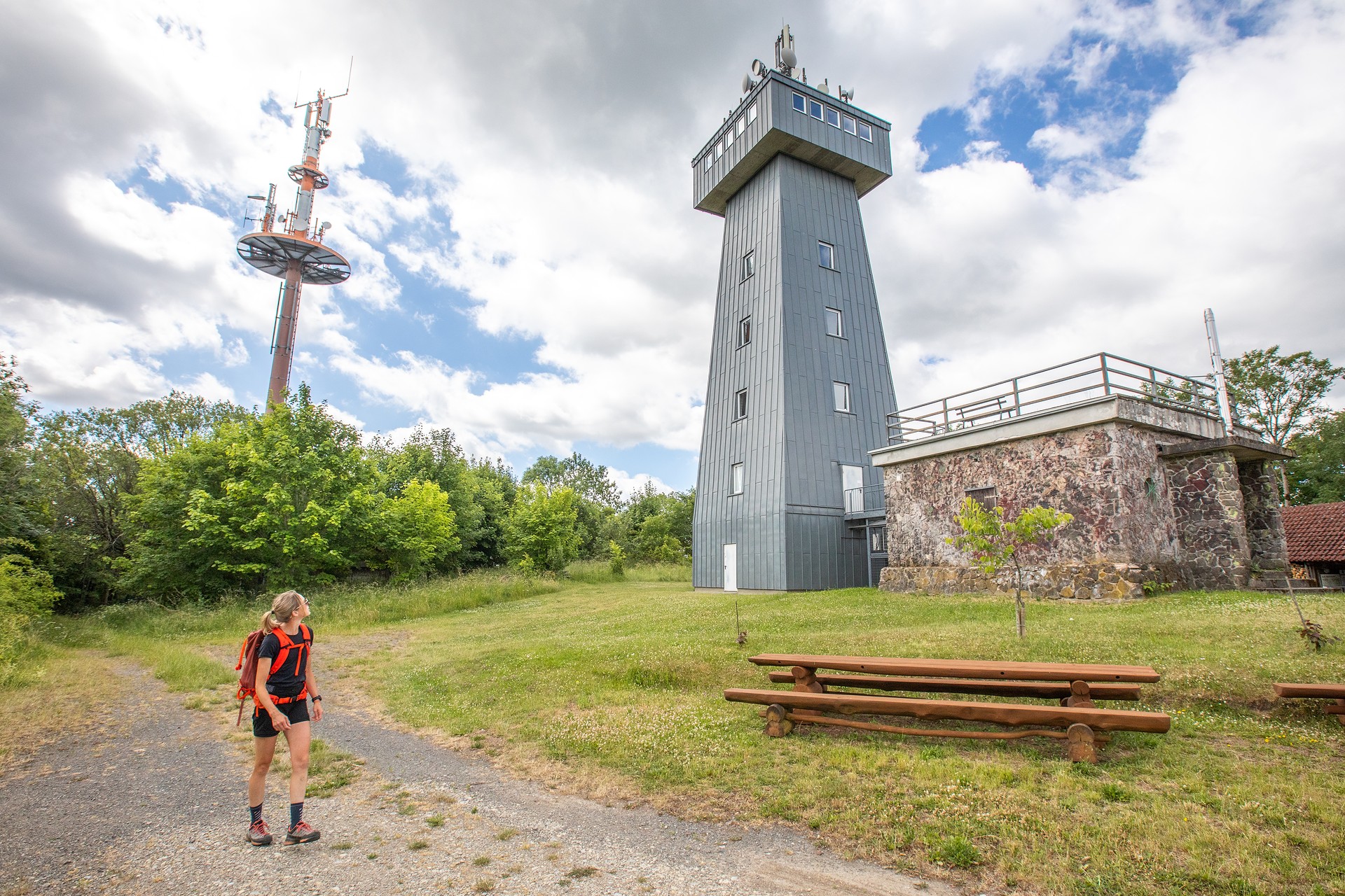 Wanderin läuft an einem grauen Aussichtsturm entlang und blickt zu diesem hoch.