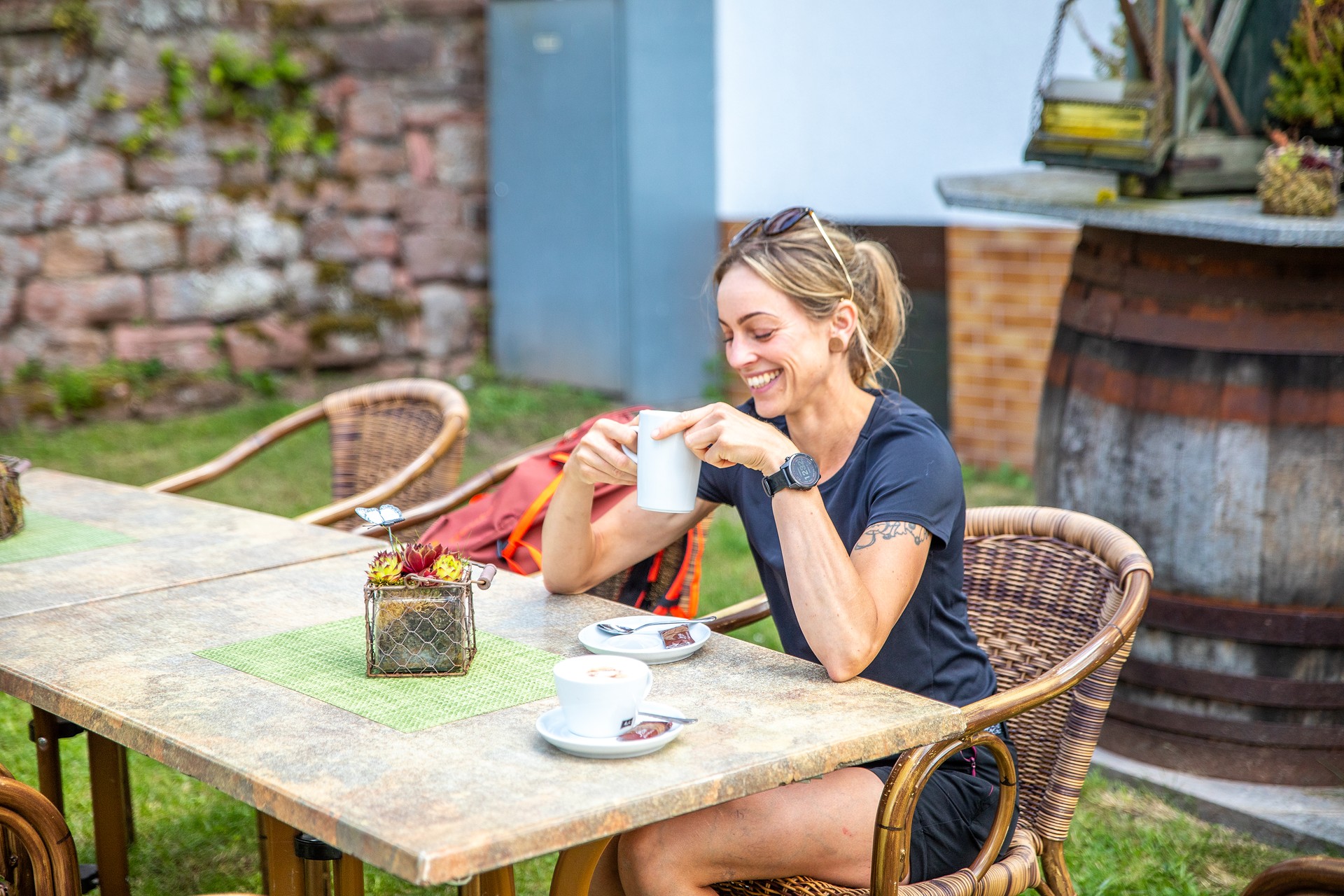 Eine Wanderin sitzt am Tisch und hält lächelnd eine Kaffeetasse.