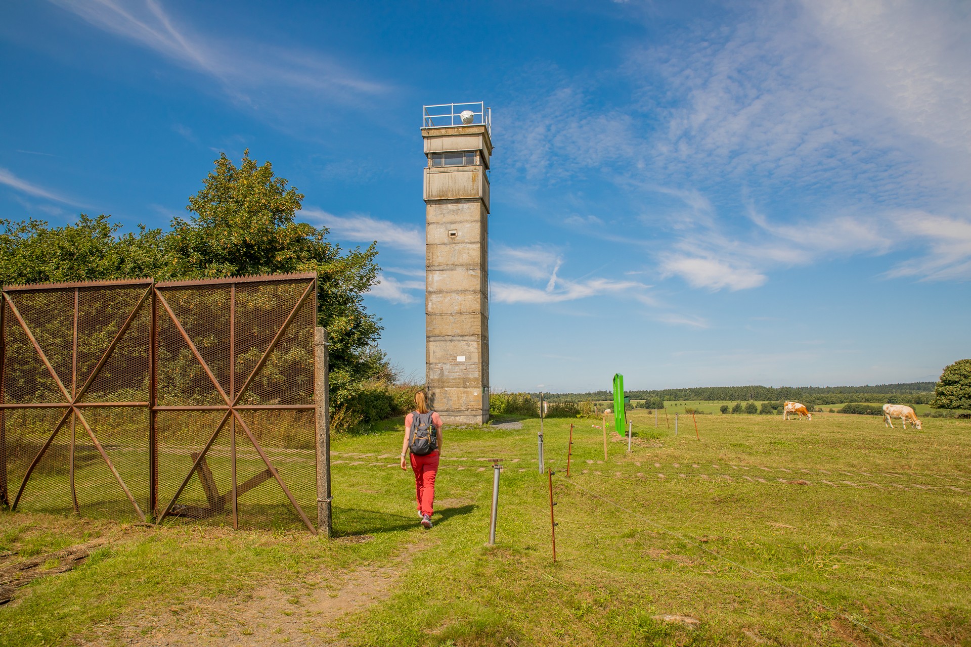 Wanderin läuft an einem ehemaligen Grenzzaun vorbei auf einen ehemaligen Grenzturm zu.