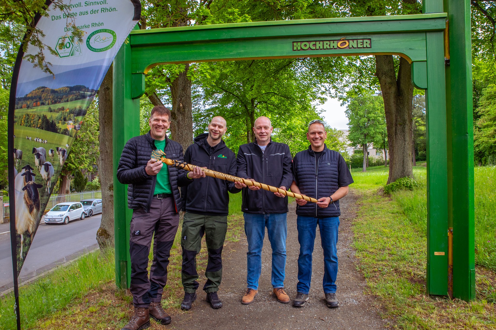 Vier Personen halten gemeinsam einen Wanderstock und stehen unter einem Holztor mit der Aufrschrift HOCHRHÖNER