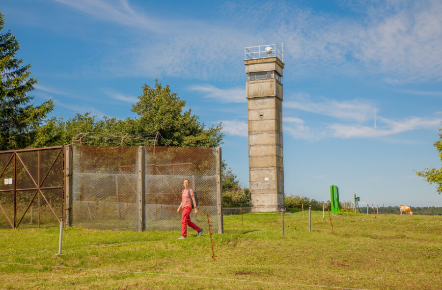 Wanderin läuft an einem ehemaligen Grenzzaun vorbei auf einen ehemaligen Grenzturm zu.