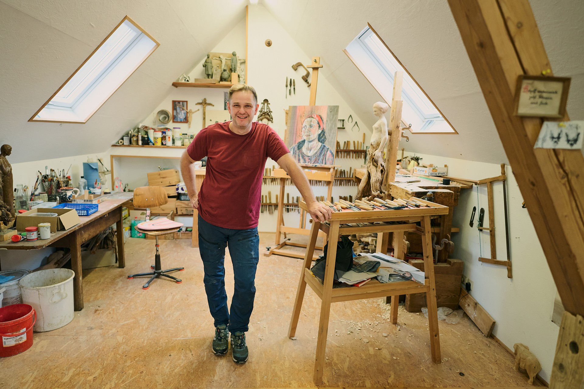 Holzschnitzer steht in seinem Atelier. Im Atelier sind viele Utensielien für Holzkunst und Malerei.