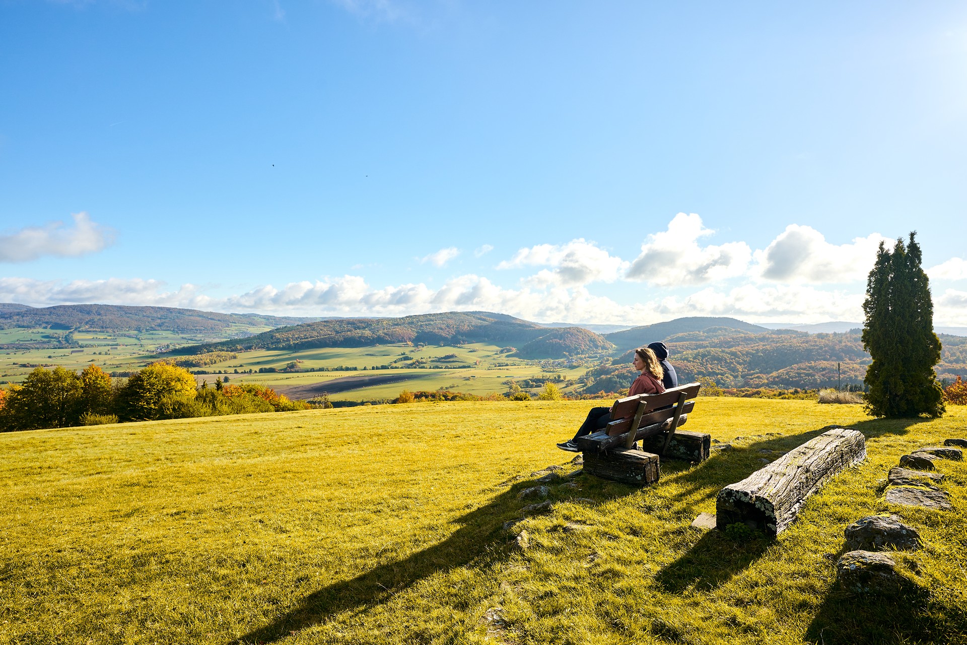 Zwei Personen sitzen auf einer Bank und blicken in die Landschaft. Im Hintrgrund ist die hügelige Rhön zu sehen.