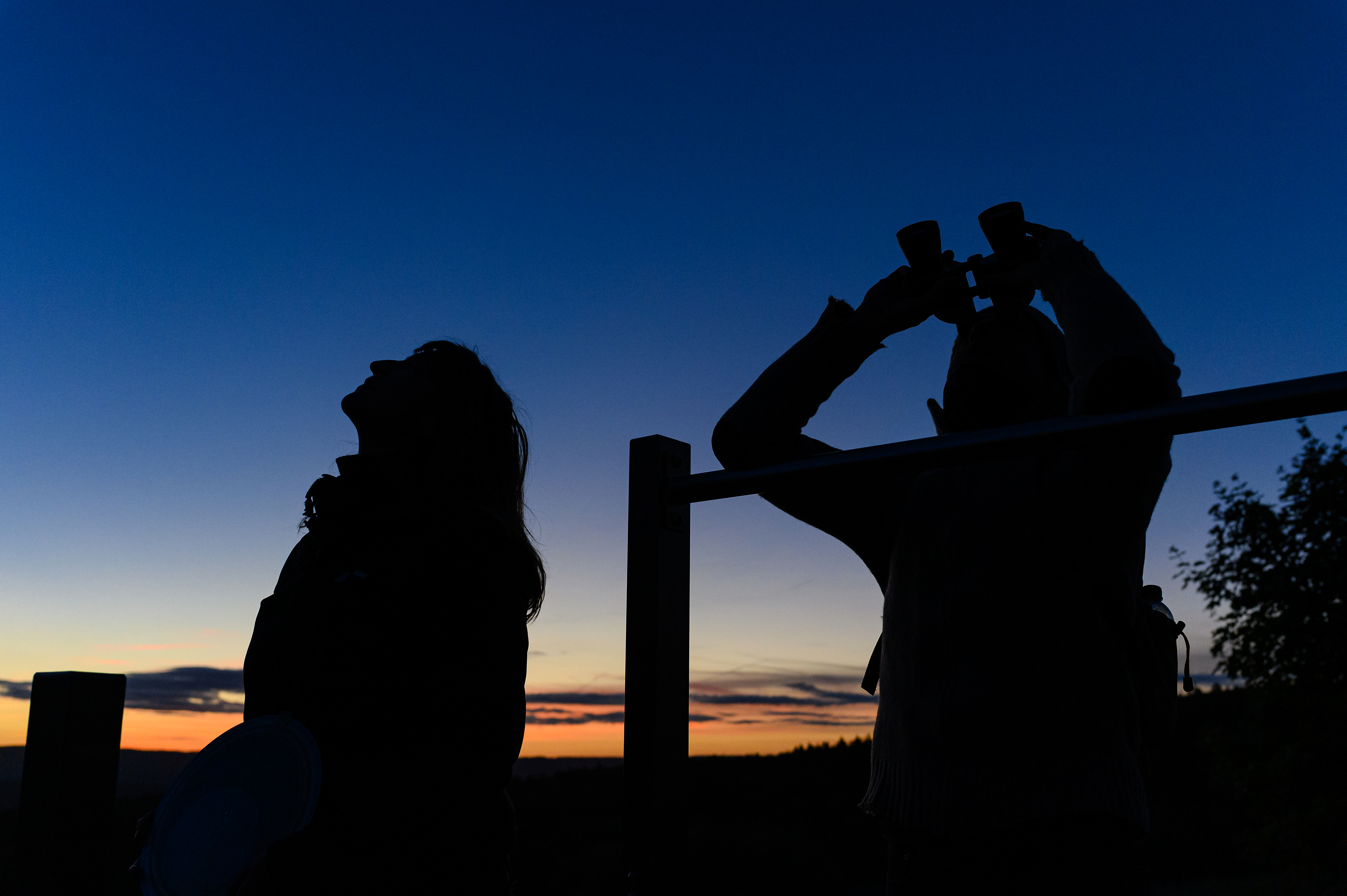 Silhouetten von zwei Personen, die in bei Nacht in den Himmel schauen. Die rechte Person hält ein Fernglas.